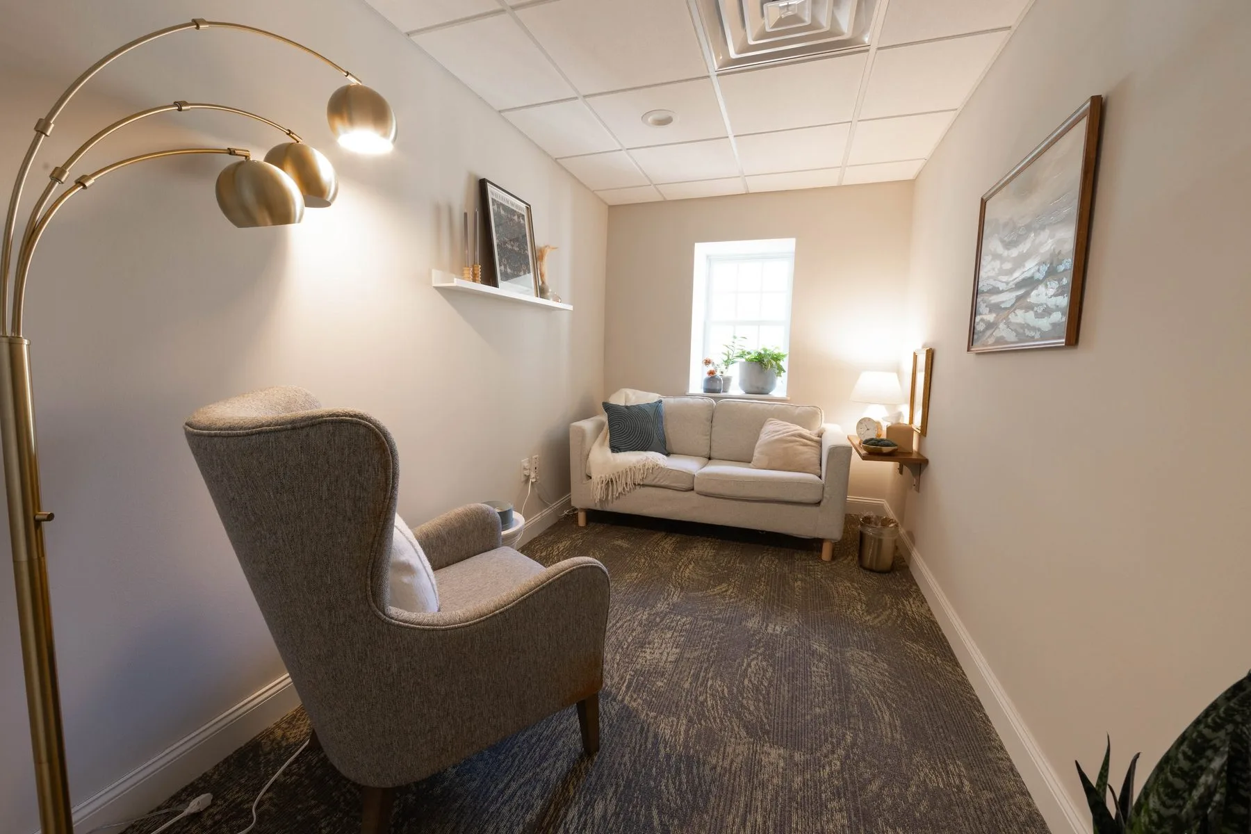 Second therapy room at the Doylestown office, with a brass arc floor lamp, a high-backed gray chair and a soft cream loveseat.